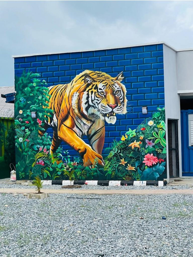 Tiger Mural by Cedar Creative Art showing a real-life looking huge tiger on a blue walled amidst beautiful flowers, giving a paradise feels.