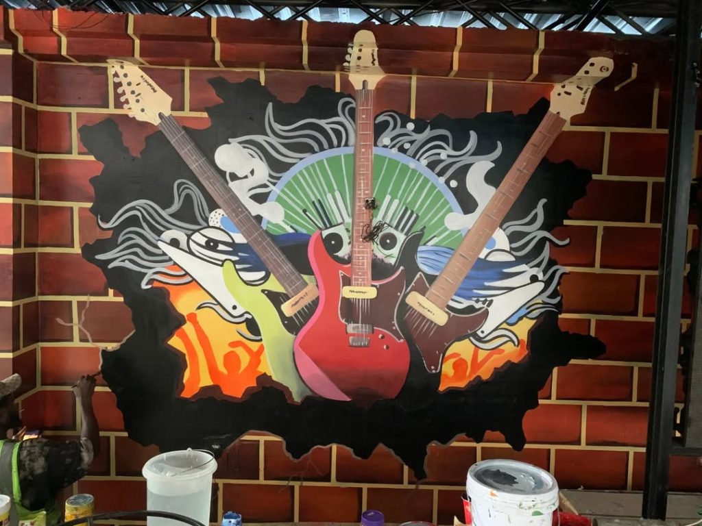 Vibrant wall mural featuring three electric guitars bursting through a brick wall, surrounded by abstract musical waves, fire elements, and urban graffiti-style art.